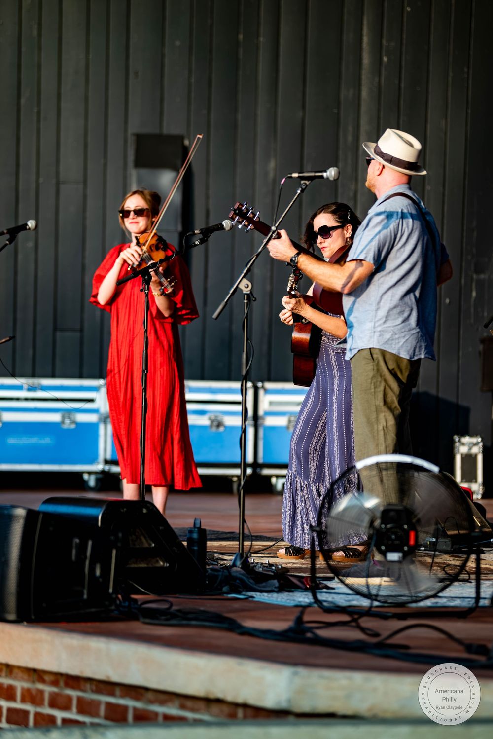 Three musicians performing on an outdoor stage, featuring a violinist in red and guitarist in blue.