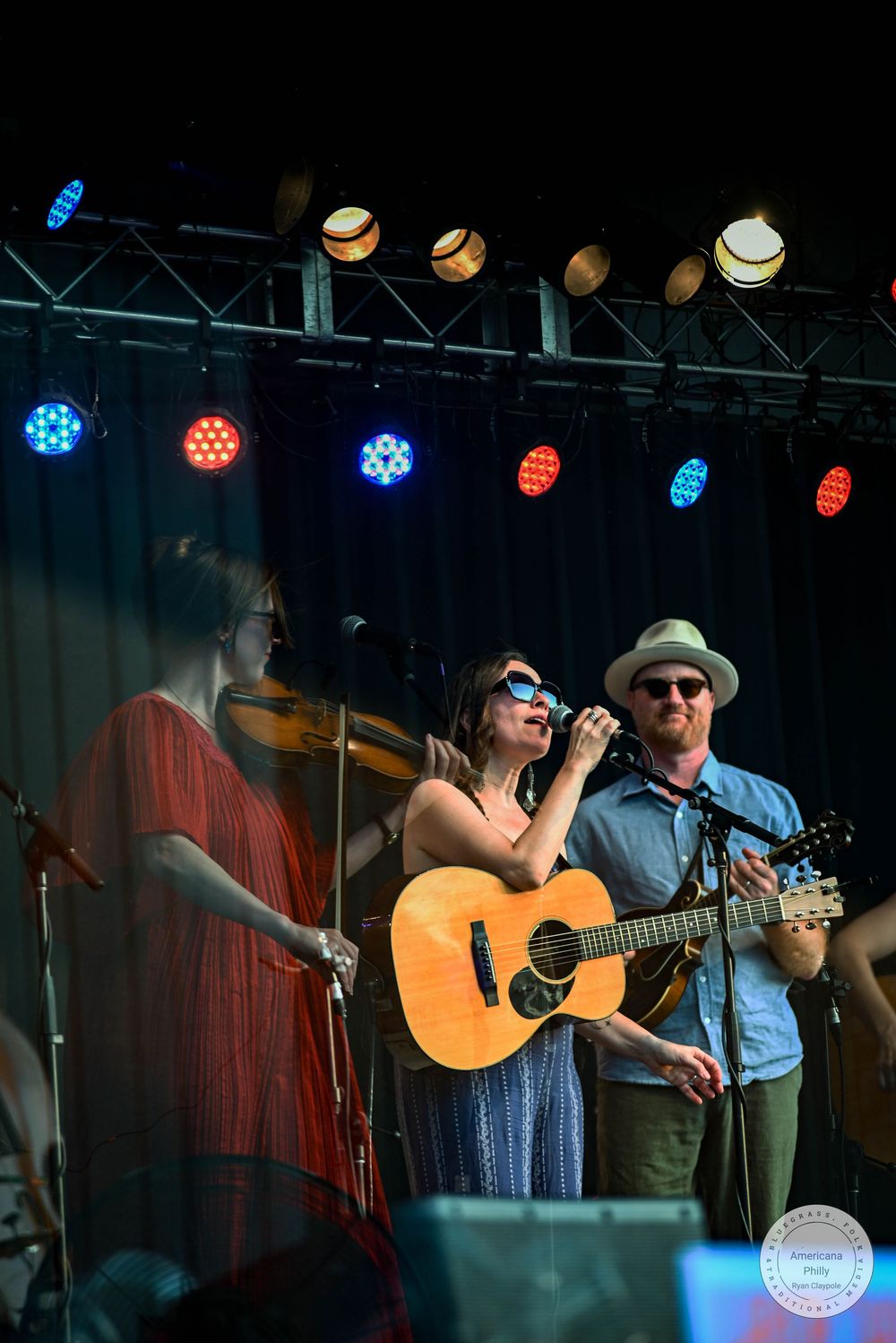 Musicians perform on stage with colorful lights, featuring a guitarist, singer, and violinist at an outdoor concert.