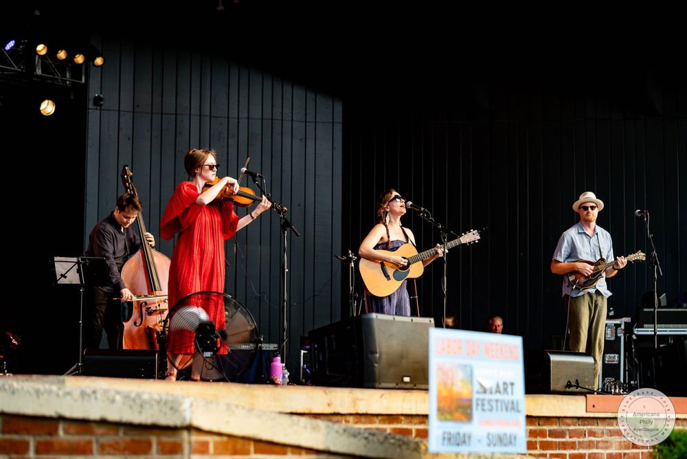 Folk band performs on outdoor stage, featuring violin, guitar, and bass at an arts festival.