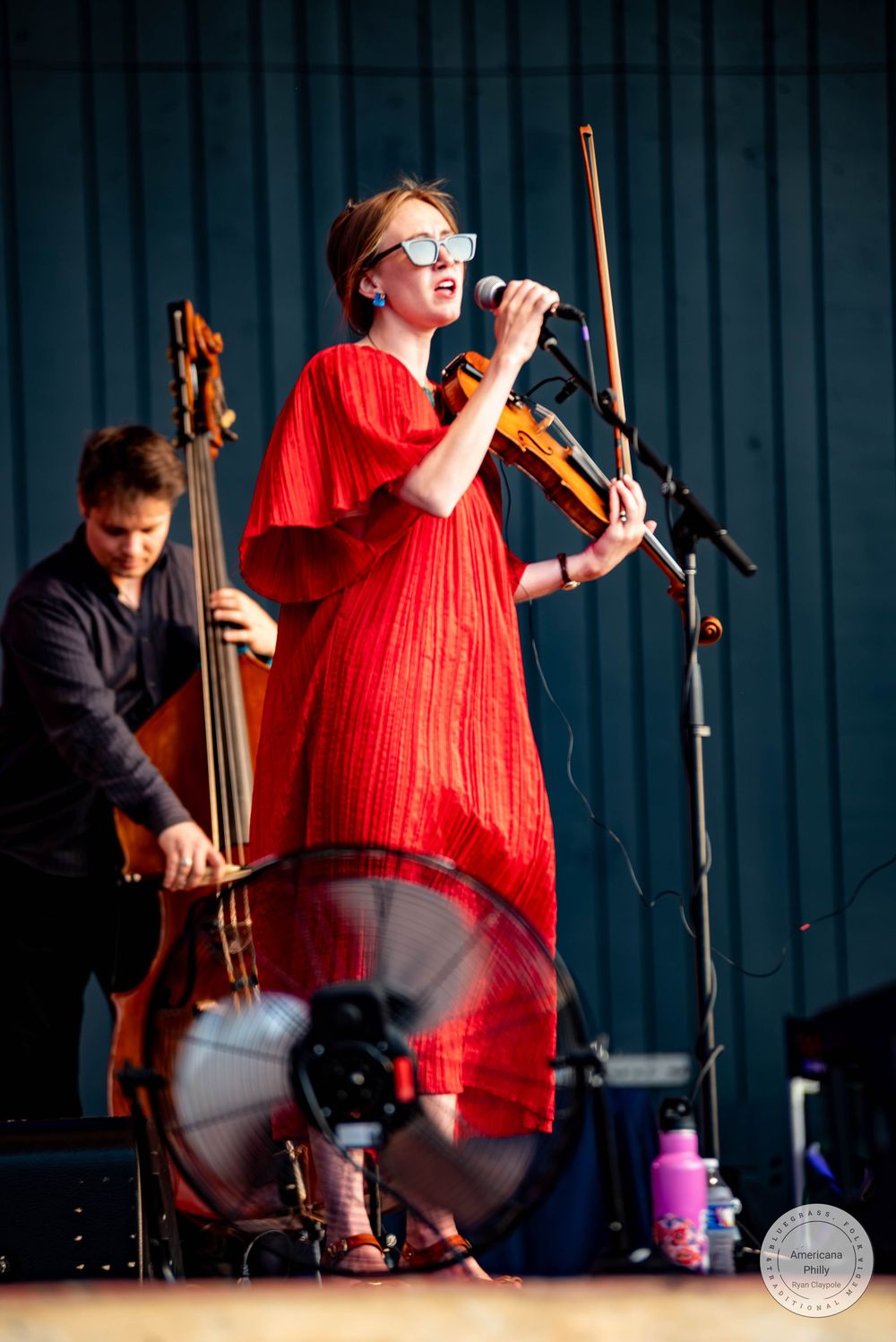 Woman in red dress plays violin and sings into microphone on stage, with bassist performing behind her.