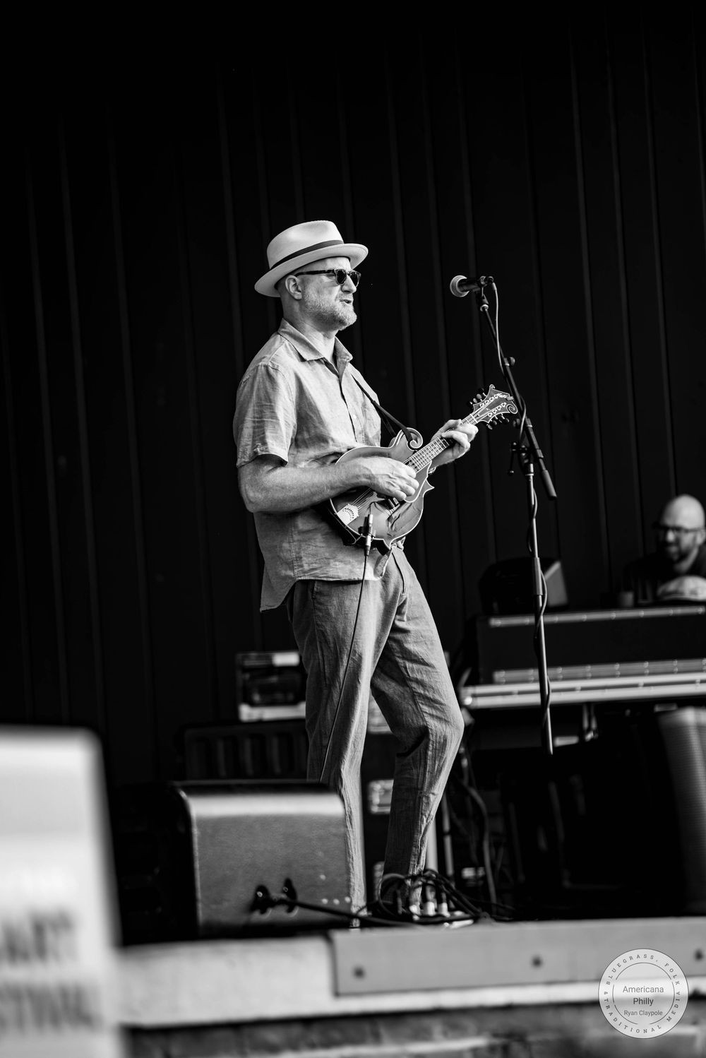 Black and white photo of a musician playing mandolin on stage wearing a fedora hat and sunglasses.