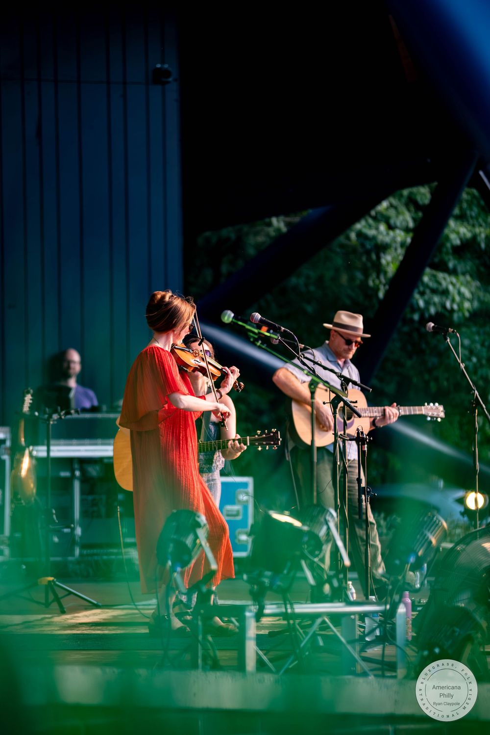 Folk duo performing on an outdoor stage at dusk, bathed in green and blue stage lighting.