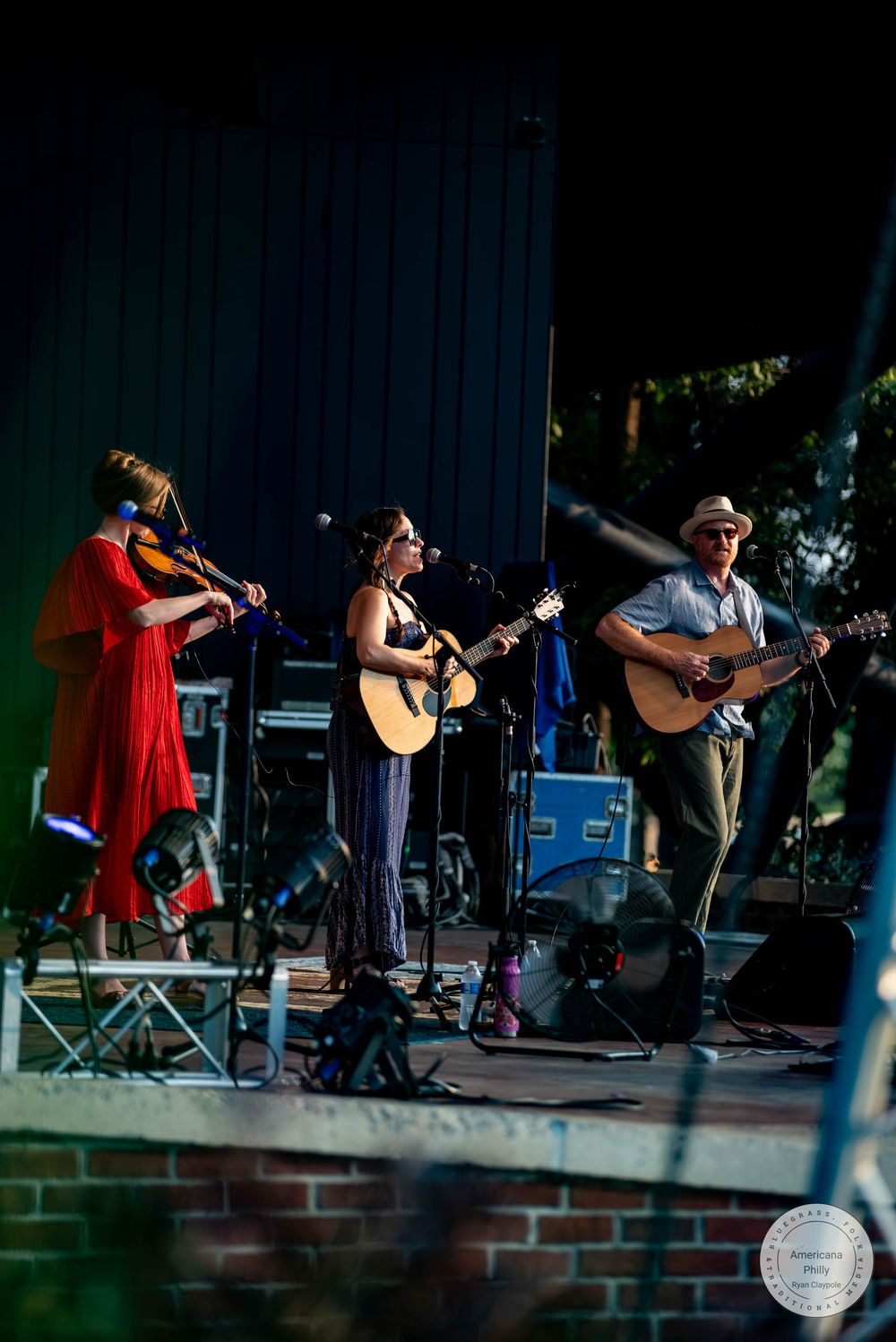 Three musicians perform outdoors on stage, with a violinist, acoustic guitarist, and singer under evening lights.