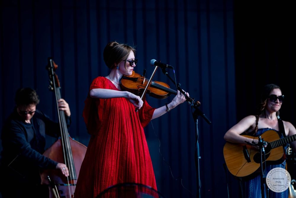 Three musicians performing live, woman in red dress playing violin with bassist and guitarist.