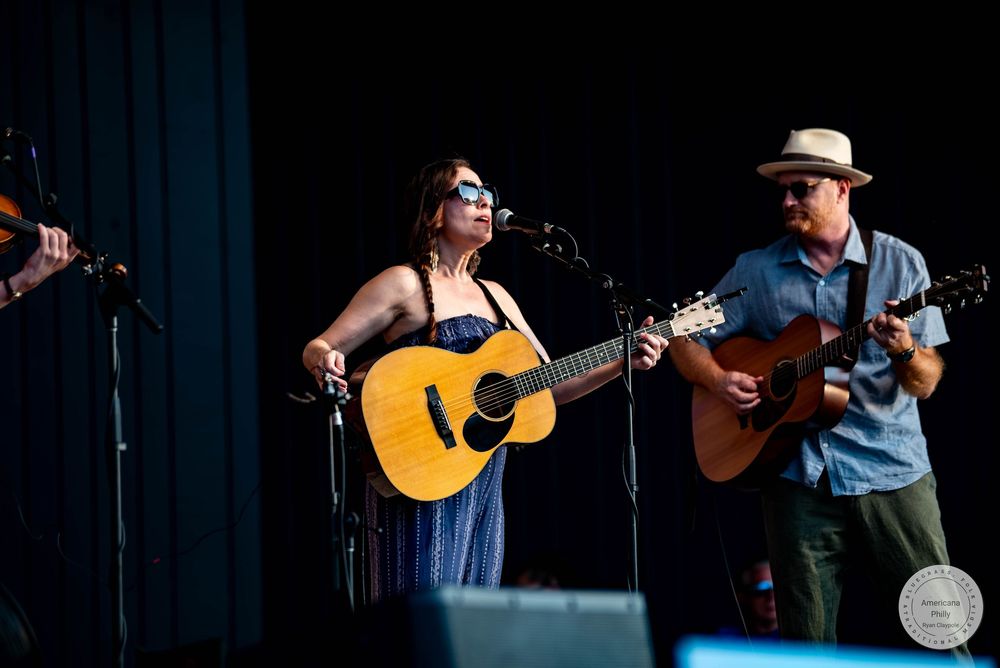 Female vocalist playing acoustic guitar on stage with a male guitarist wearing a straw hat.