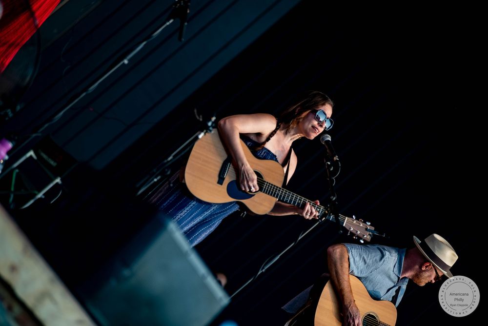 Female guitarist performing on a dimly lit stage with a male guitarist in a straw hat nearby.