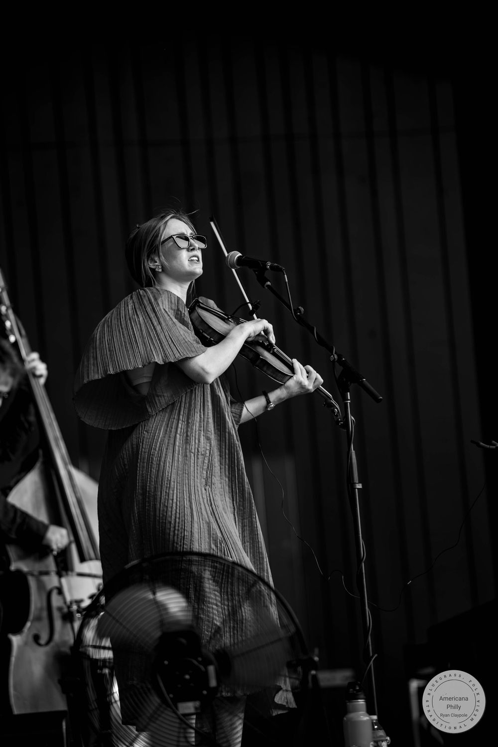 Black and white photo of a woman playing violin on stage near a microphone during a live performance.