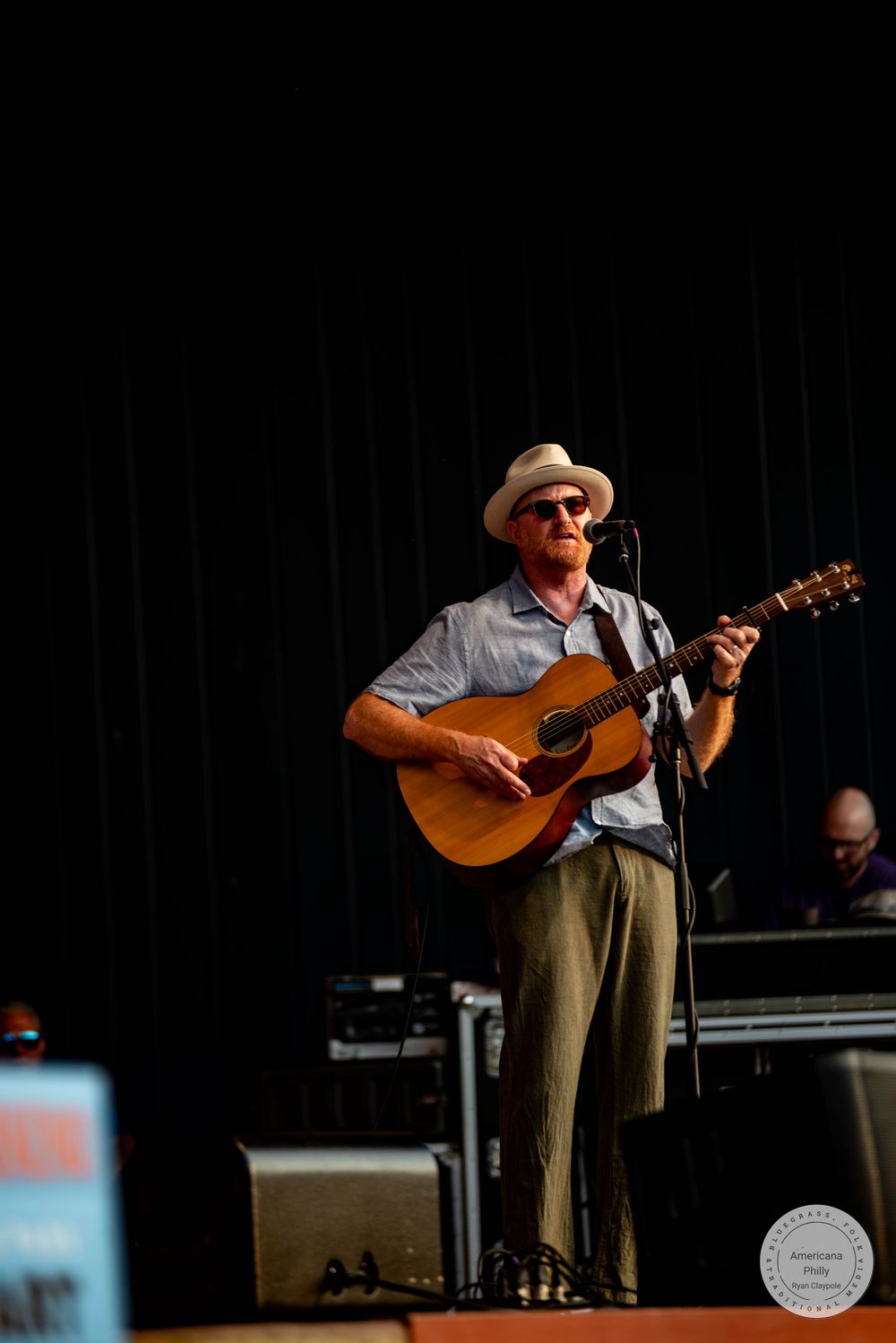 Musician in white hat playing acoustic guitar on stage at an outdoor music festival.