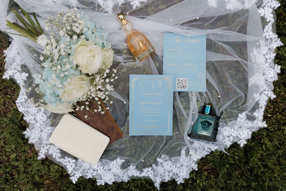 Elegant wedding flat lay with blue invitation suite, bridal bouquet of white roses and baby's breath, perfume, and veil on grass.
