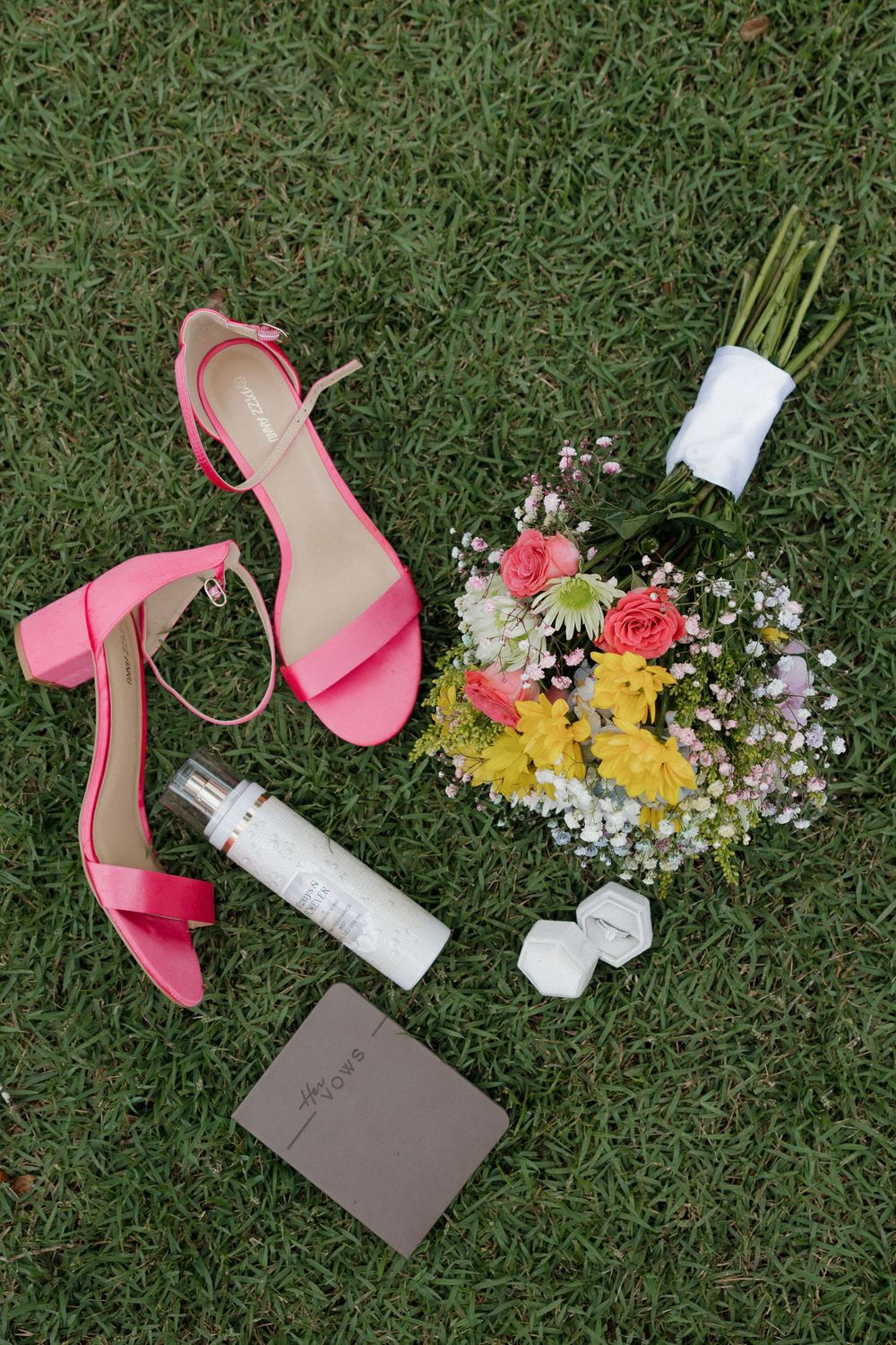 Pink block heel sandals, floral bouquet, perfume bottle, and ring box arranged on green grass for a wedding flat lay.