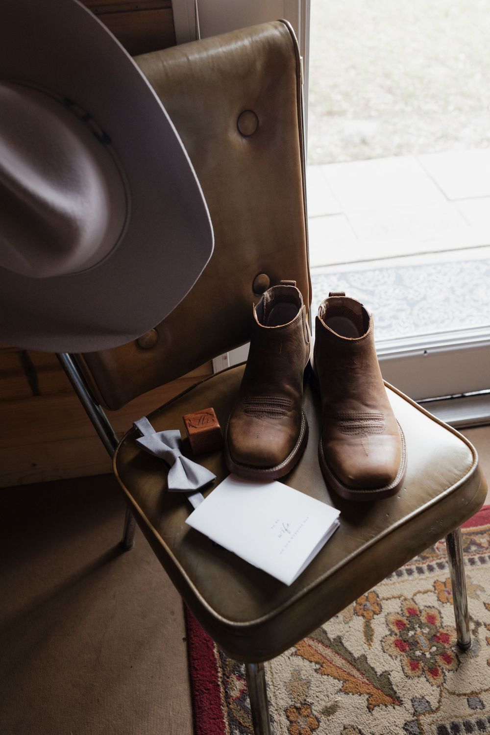 Brown leather cowboy boots, gray felt hat, bow tie, and wedding cards arranged on a vintage metal chair near a window.