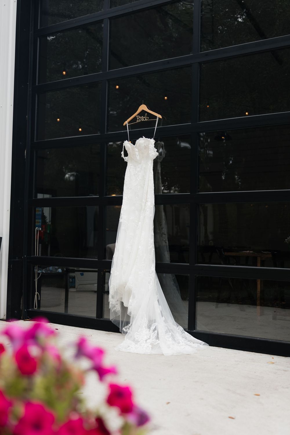 White lace wedding dress hanging on a wooden hanger against black glass windows with pink flowers in foreground.