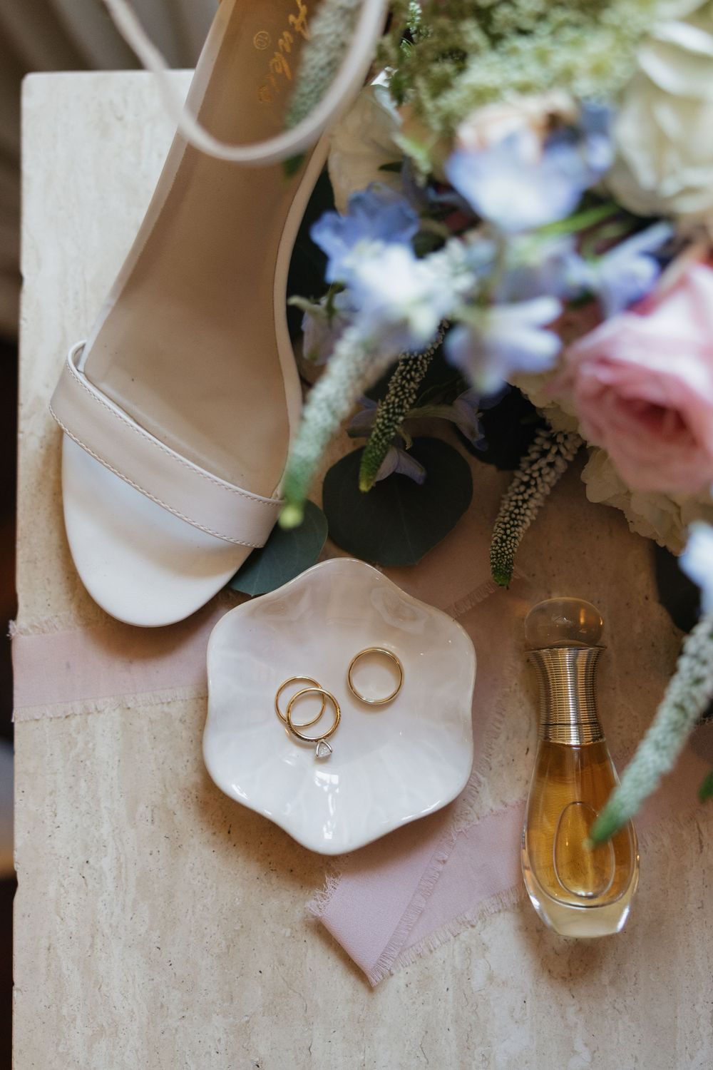Wedding rings on a white dish beside perfume and bridal heels with a floral bouquet.