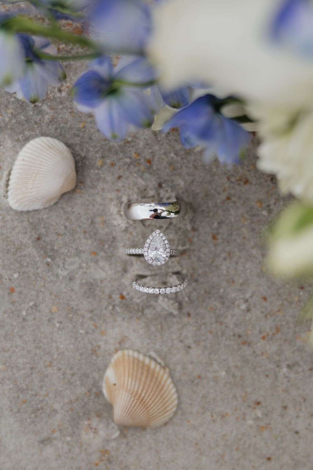 Beach wedding rings with pear diamond engagement ring on sand with seashells and blue flowers.