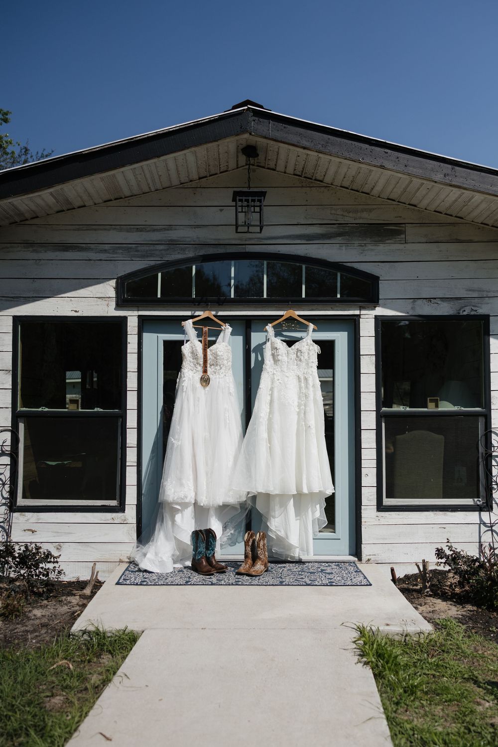 Two white wedding dresses hanging on a rustic white farmhouse door with cowboy boots below on a sunny day.