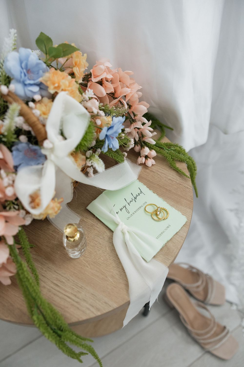 Bridal flat lay with pastel flower bouquet, gold wedding rings, perfume, and nude heels on a wooden stand.