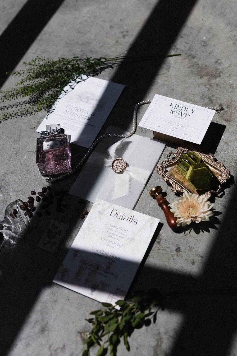 Elegant wedding invitation suite with perfume bottles, wax seal necklace, and florals on concrete surface.