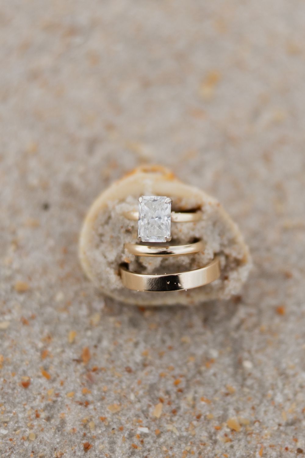 Gold wedding band and radiant-cut diamond engagement ring resting on a seashell in sandy beach setting.