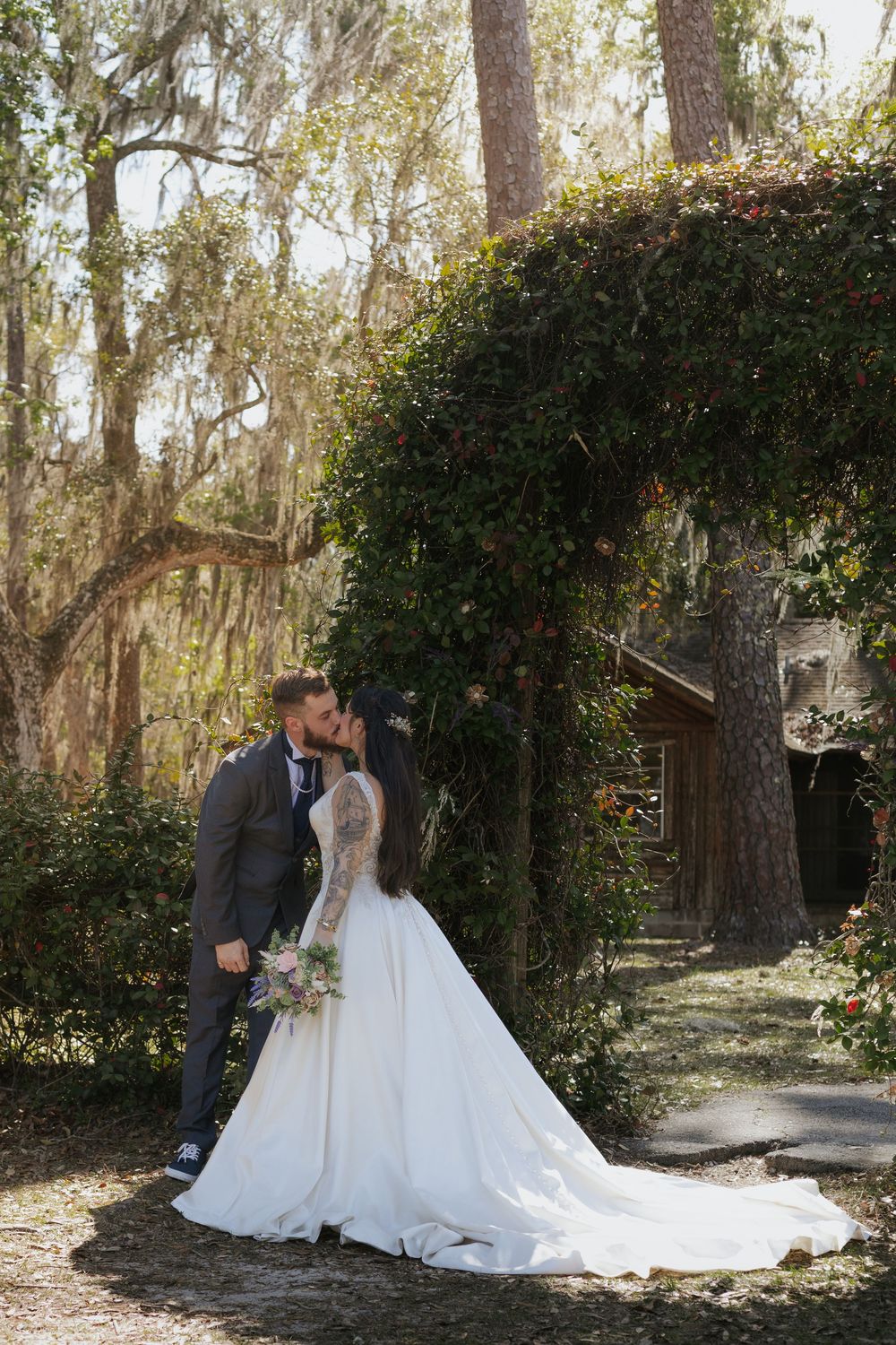 Bride and groom share a romantic kiss under ivy-covered arch surrounded by moss-draped trees at outdoor wedding.