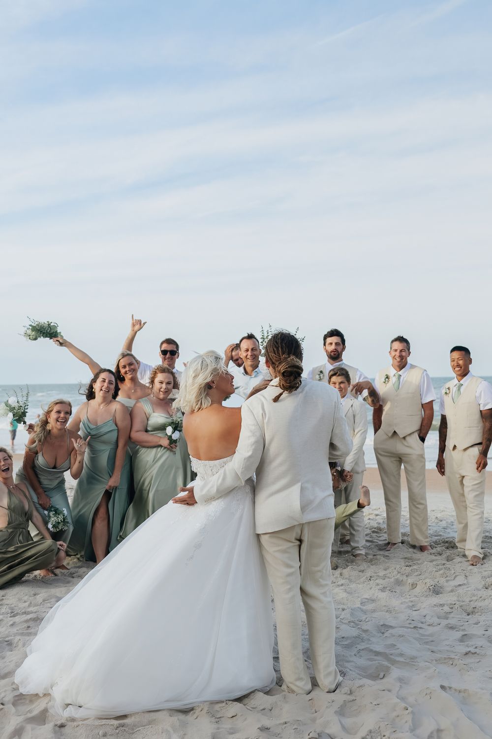 Bride and groom kiss on beach surrounded by cheering wedding party in sage green and tan attire.