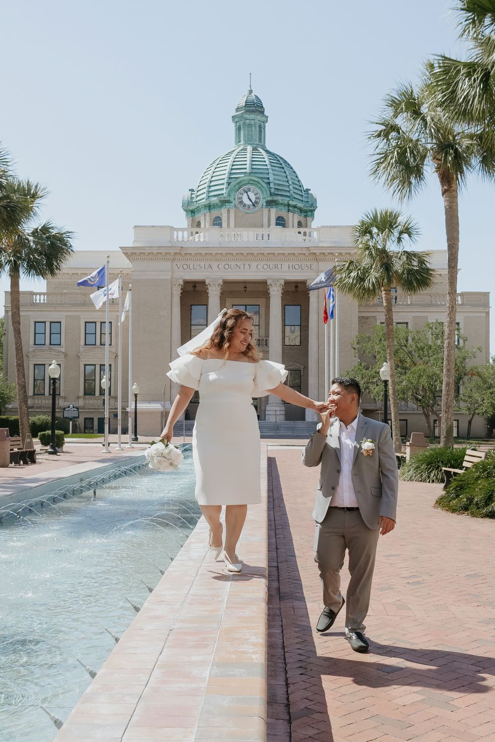 Newlyweds walk hand-in-hand near fountain outside Volusia County Courthouse on sunny wedding day in Florida.
