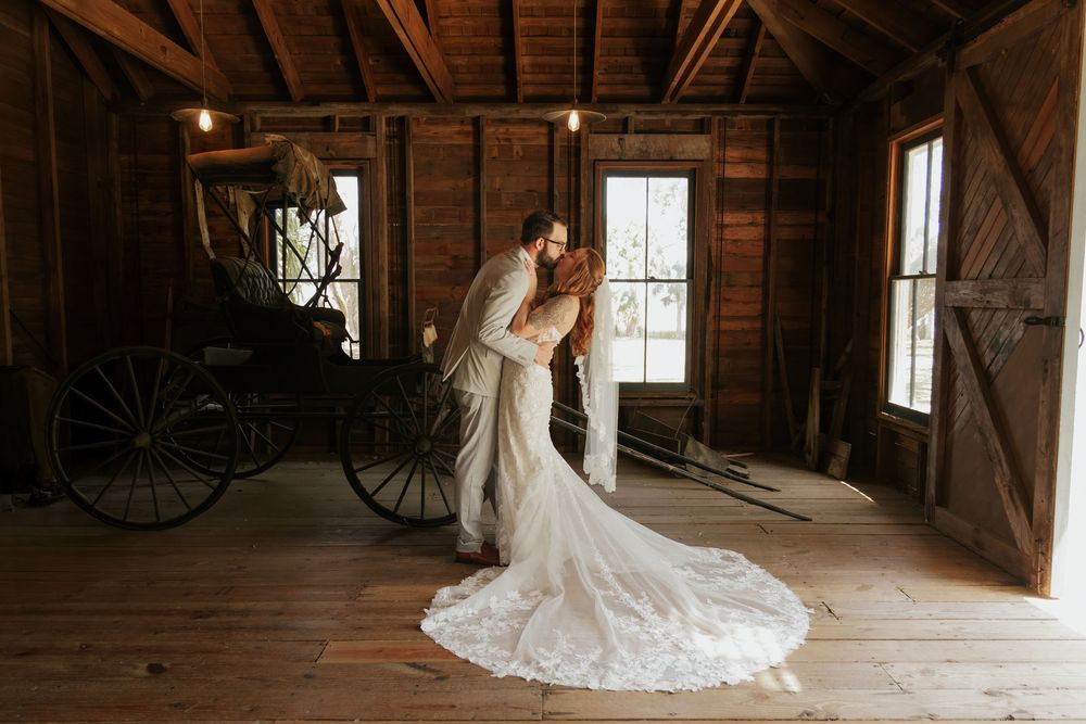 A bride and groom share a romantic kiss in a rustic wooden barn, with a vintage carriage in the background.