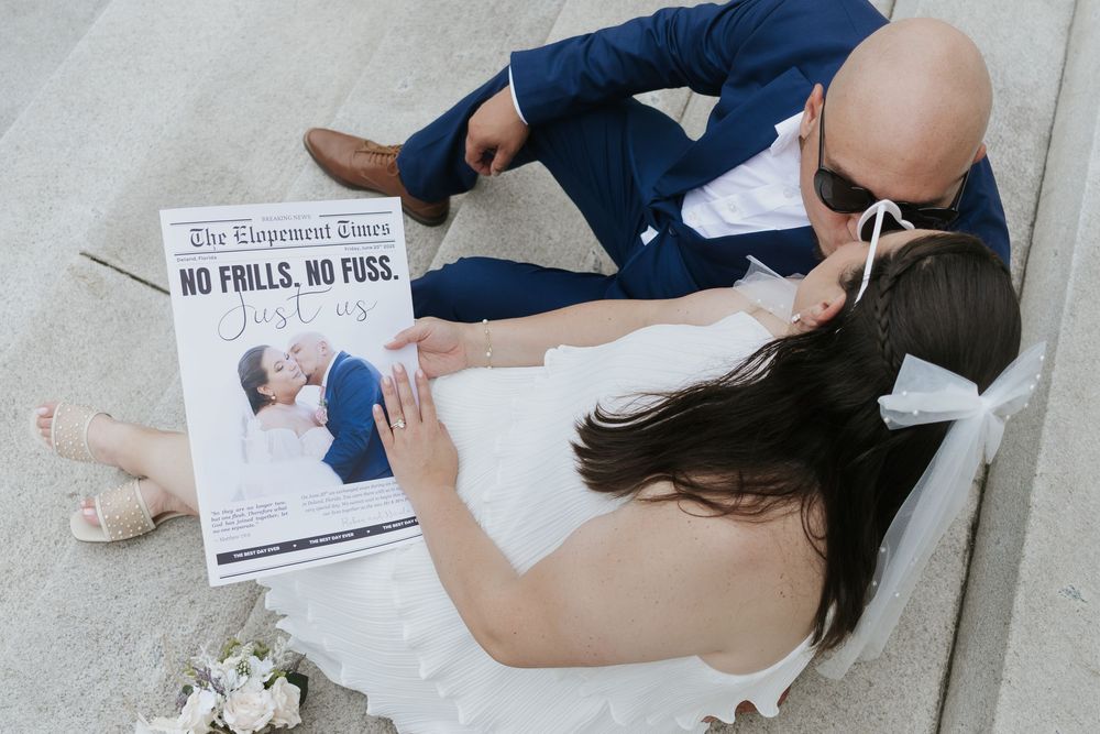 Couple in wedding attire reading The Elopement Times newspaper together on steps, bride in white dress, groom in navy suit.