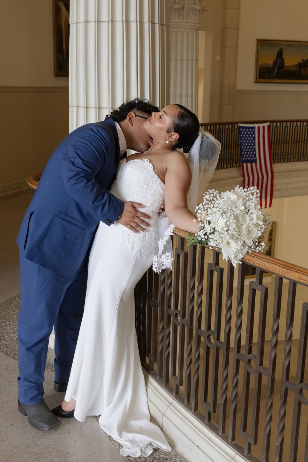 Newlywed couple sharing a kiss at city hall wedding, bride in white gown holding bouquet, groom in blue suit.