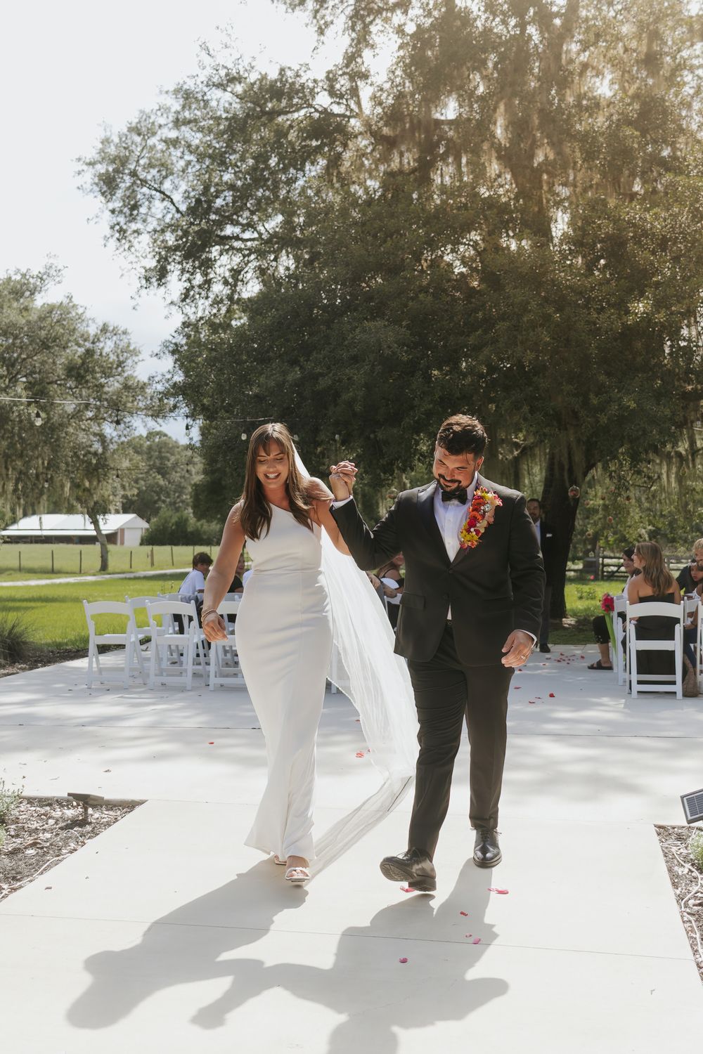 Joyful bride and groom walk down outdoor wedding aisle, bride in white gown holding veil, groom in black suit with floral lei.
