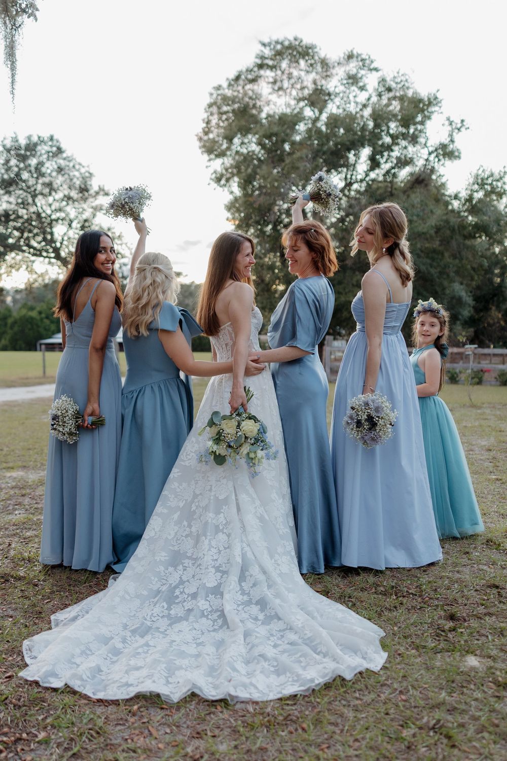 Bride in lace gown poses with bridesmaids in blue dresses and flower girl outdoors at wedding.