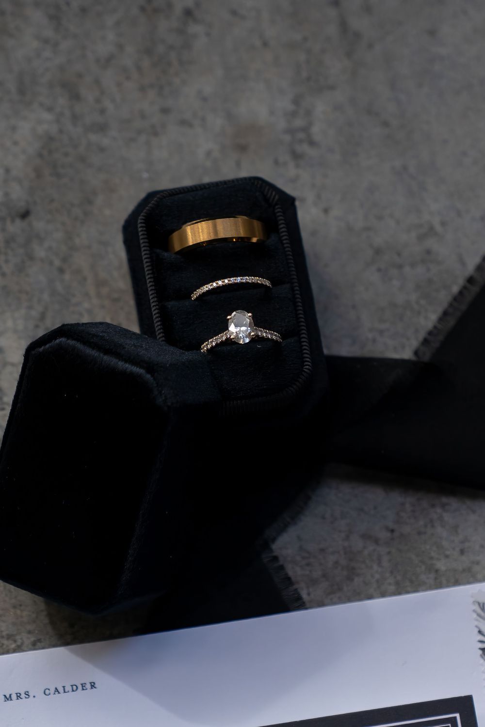 Three gold wedding rings displayed in a black velvet ring box, with a diamond solitaire engagement ring, beside Mrs. Calder stationery.