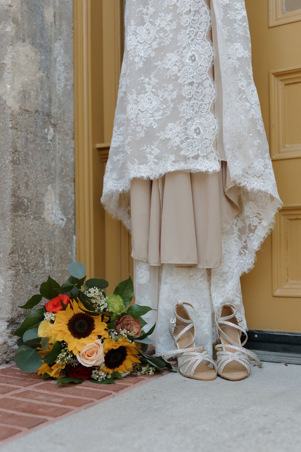 Bridal wedding dress with lace overlay, strappy heels, and sunflower bouquet displayed by a yellow door.