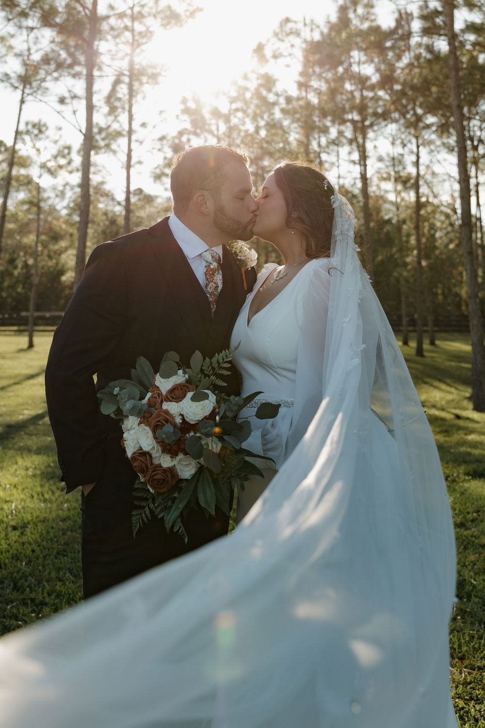 Bride and groom share a romantic kiss outdoors at golden hour, with veil flowing and bridal bouquet in hand.