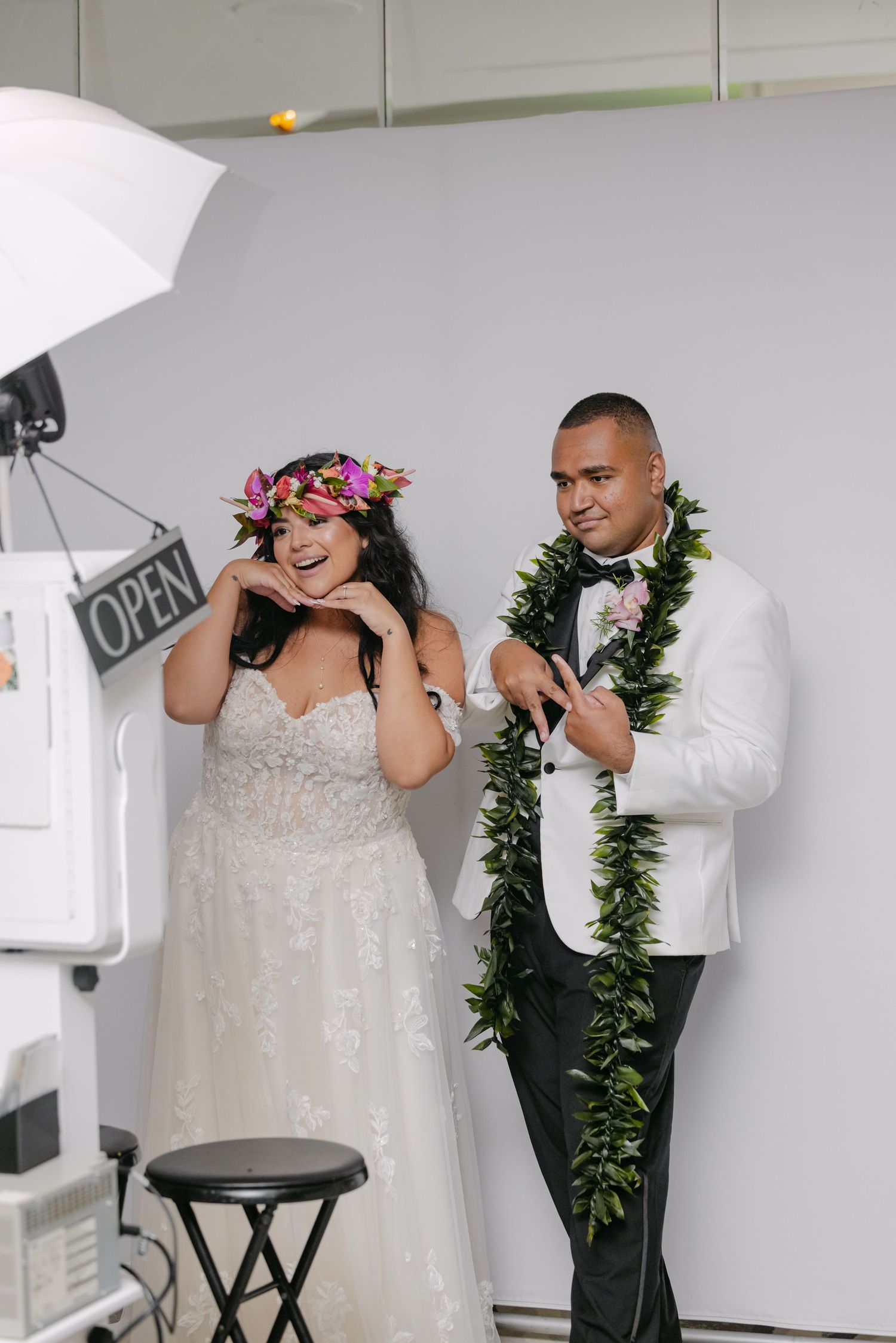 Bride in floral crown and groom in white tuxedo with green lei posing playfully at a wedding photo booth.