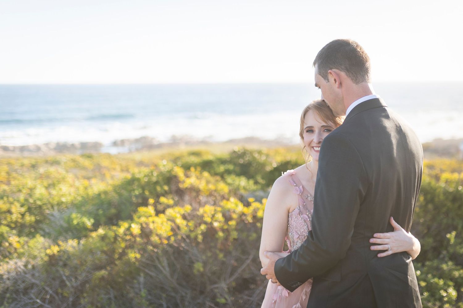 Couple embracing near coastal wildflowers at golden hour, woman in pink dress smiling over shoulder.