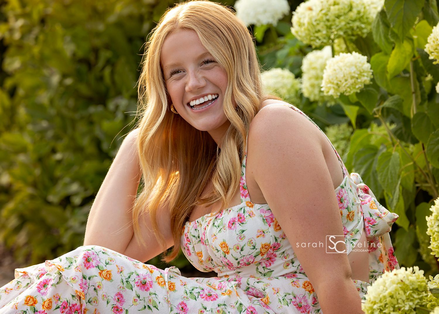 Smiling young woman in floral dress sitting among white hydrangeas in a lush green garden.