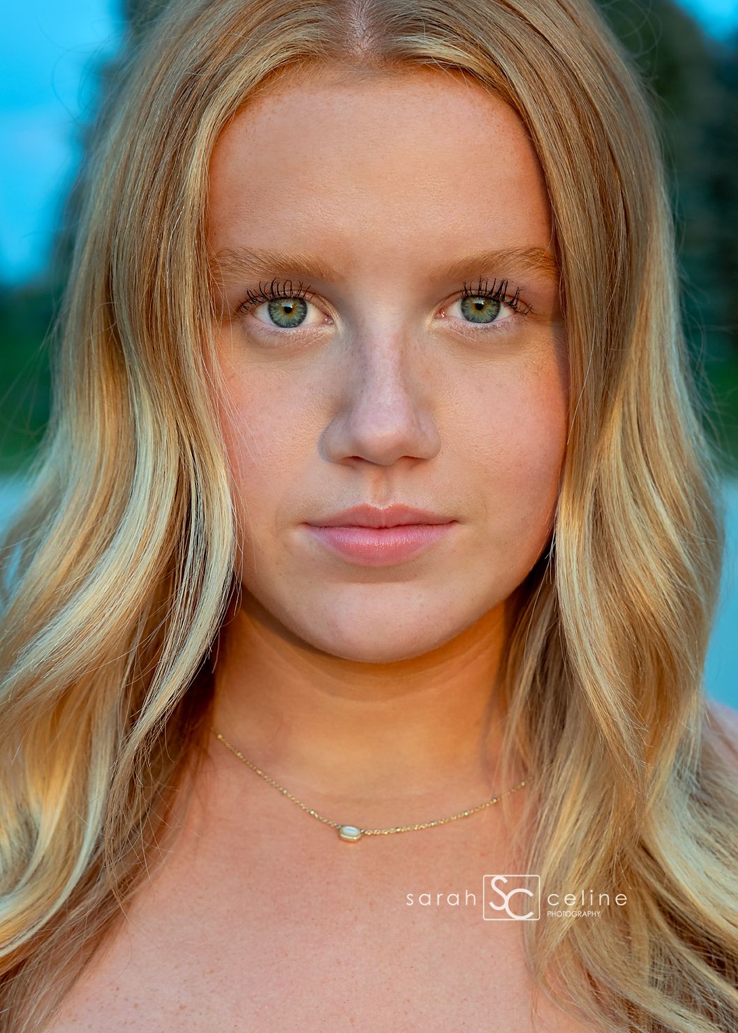 Young blonde woman with green eyes and freckles wearing a delicate gold necklace, photographed outdoors in natural light.