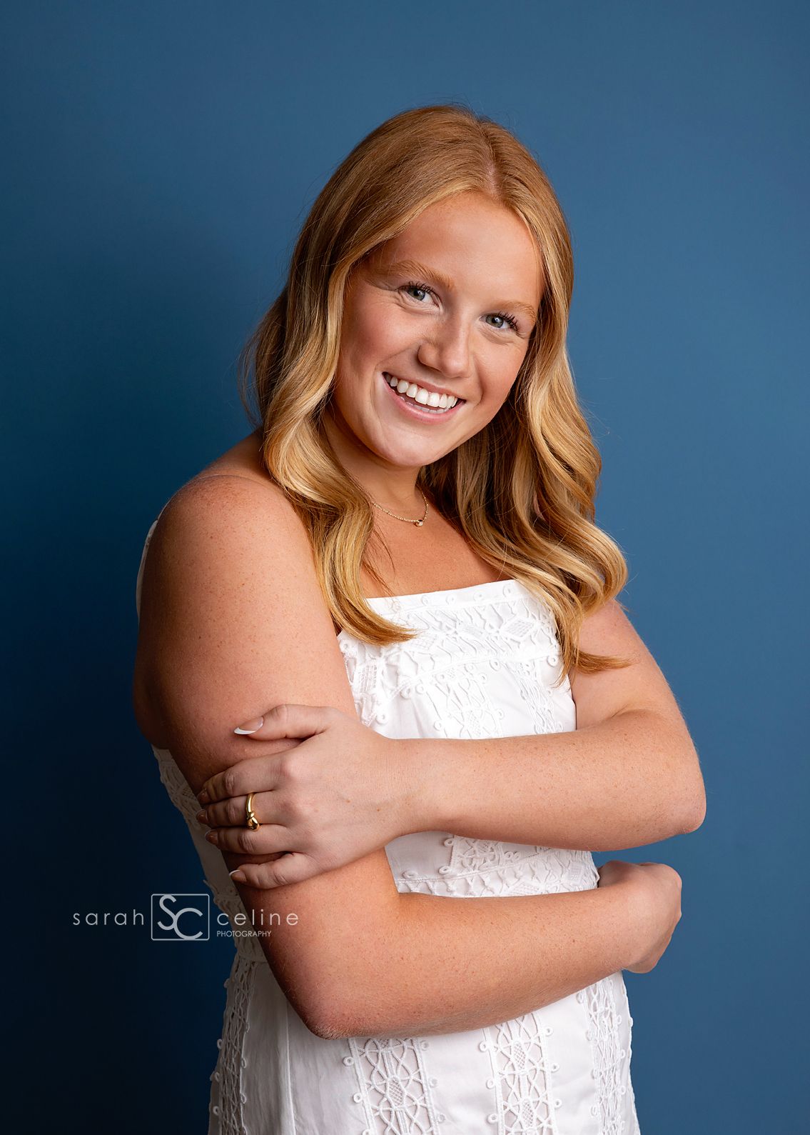 Smiling teenage girl with long blonde hair in white dress poses confidently against a blue studio backdrop.
