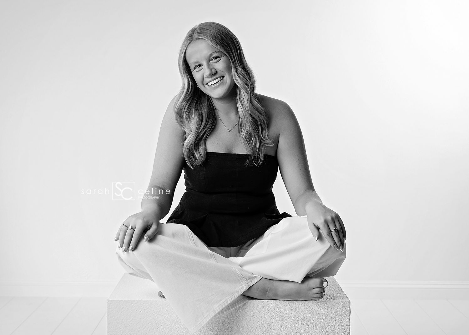 Black and white photo of a smiling young woman sitting cross-legged in a black top and white pants.