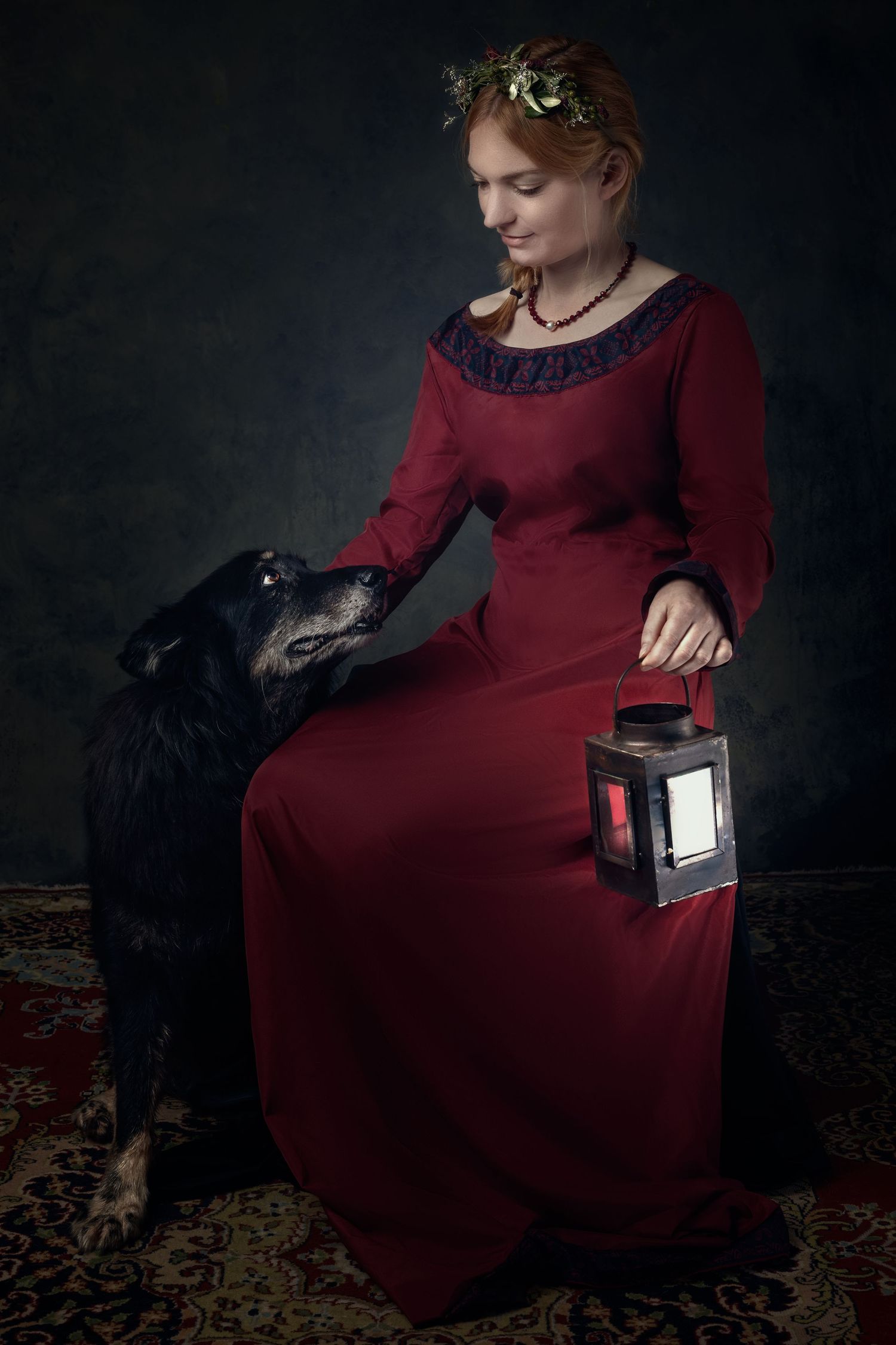 Woman in red medieval dress holds lantern beside black dog, wearing floral crown, in dramatic dark studio portrait.