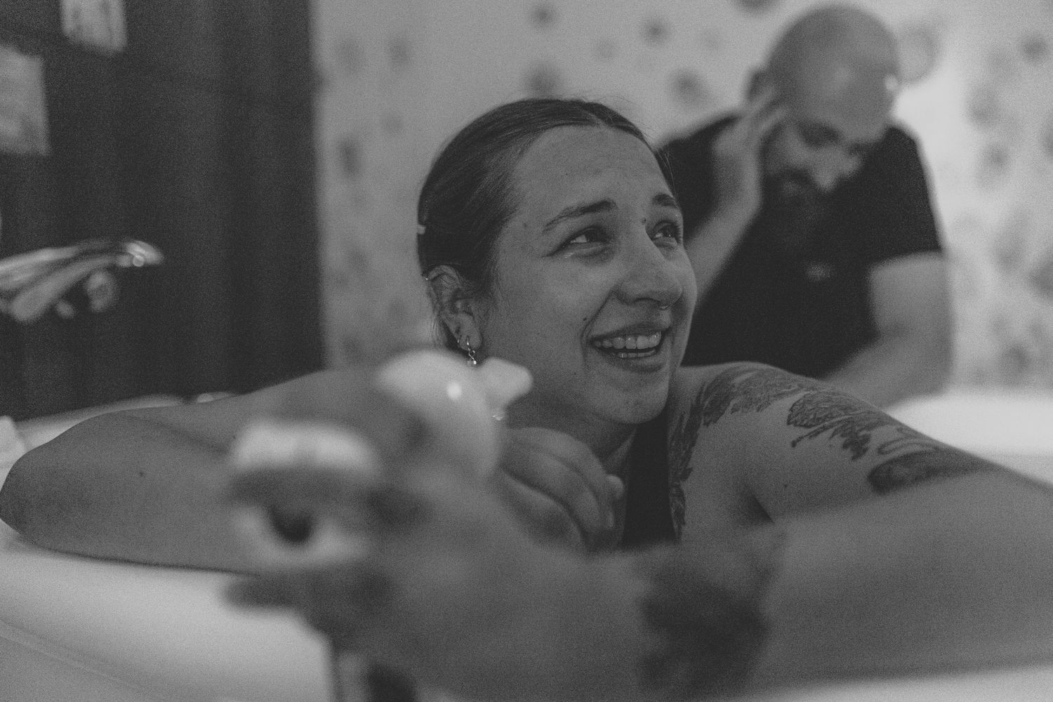 Black and white photo of a smiling tattooed woman relaxing in a bathtub with a man on phone in background.