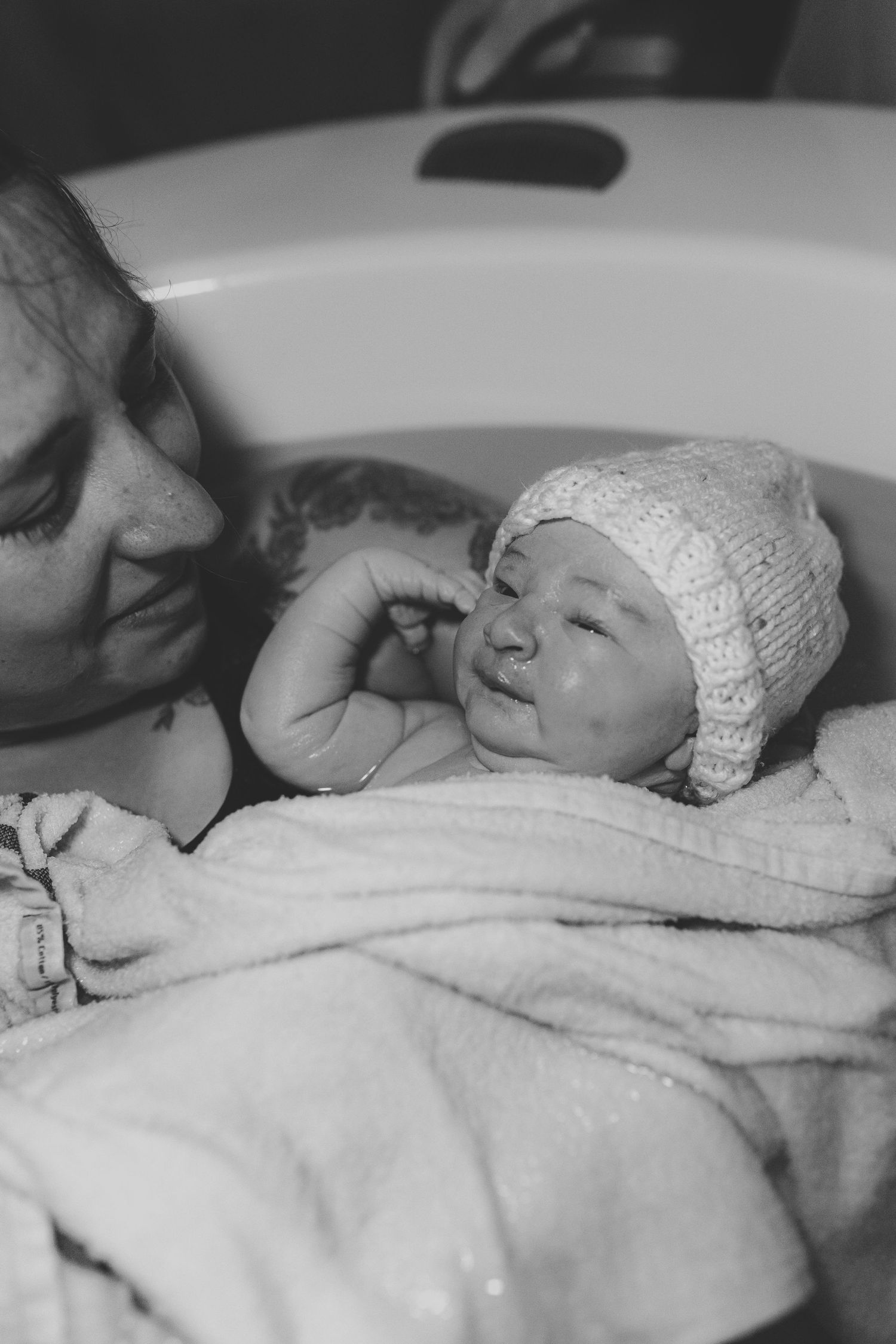 Black and white photo of a newborn baby in a knit hat wrapped in a towel, cradled by a smiling mother near a bathtub.