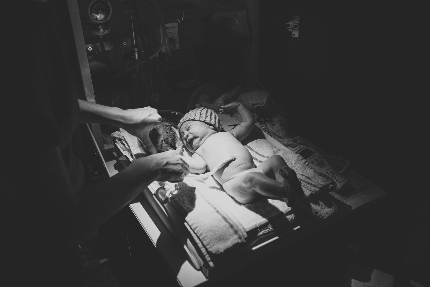 Black and white photo of a newborn baby lying in a hospital bassinet under warm light with a caregiver's hand nearby.