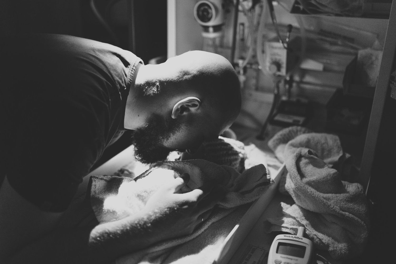 Black and white photo of a bald man tenderly kissing a newborn baby wrapped in white blankets in a hospital.