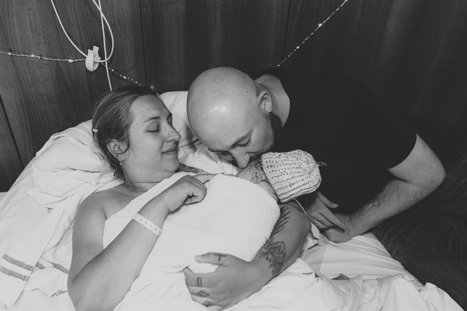 Black and white photo of new parents in hospital bed lovingly kissing their newborn baby after birth.