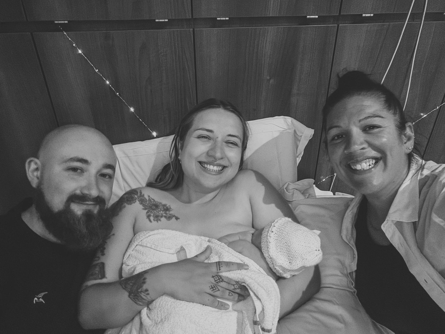 Black and white photo of new parents smiling in hospital bed holding newborn baby with support person beside them.