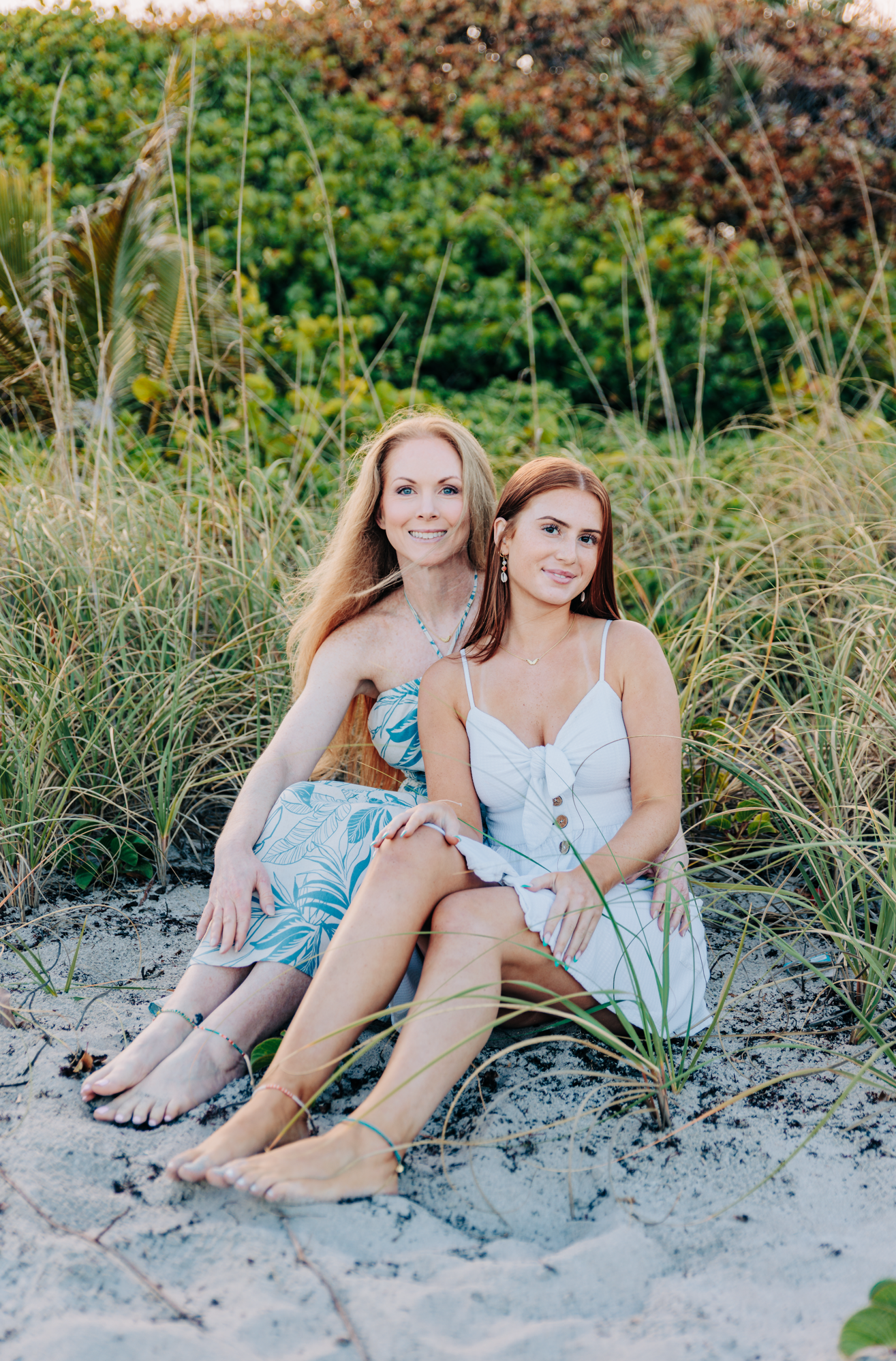 Two women in summer dresses sit together on sandy beach dunes surrounded by lush green sea grass.