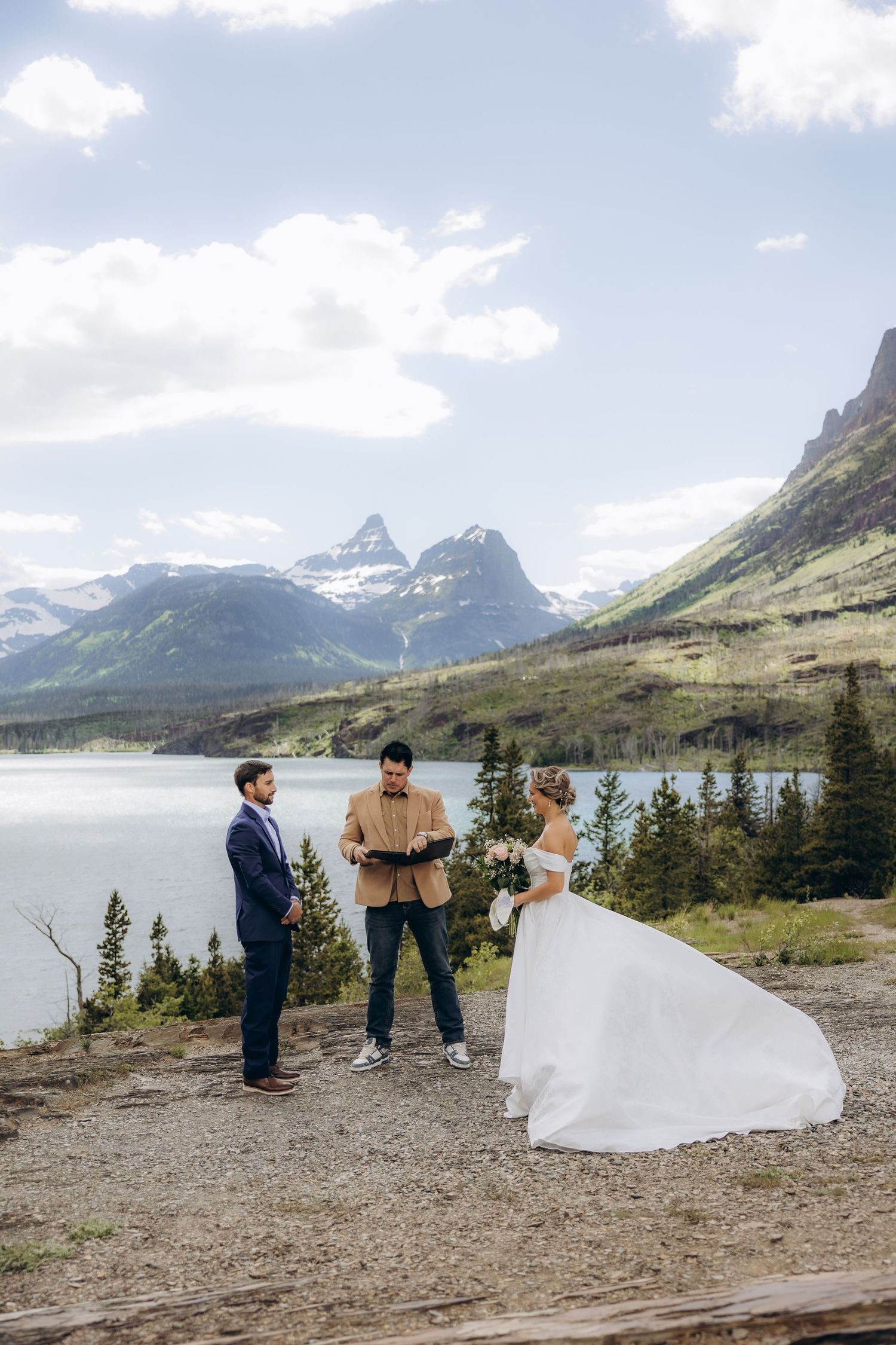Outdoor elopement ceremony with bride and groom facing officiant, mountain lake and snow-capped peaks backdrop, Glacier National Park.