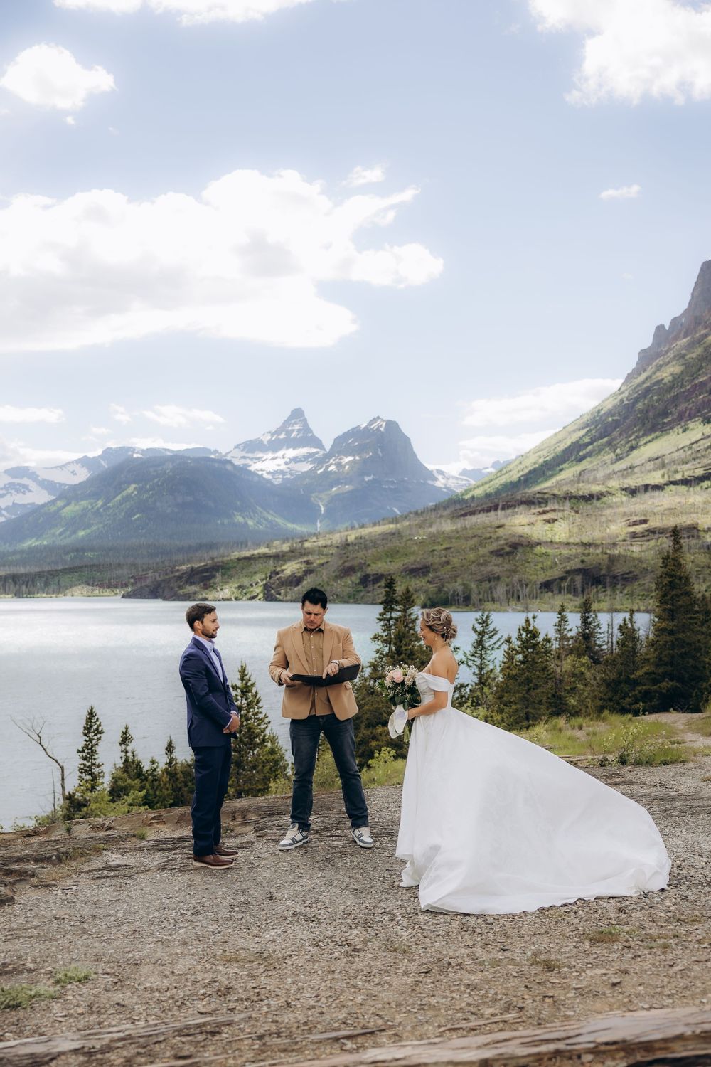Outdoor elopement ceremony with bride and groom facing officiant, mountain lake and snow-capped peaks backdrop, Glacier National Park.