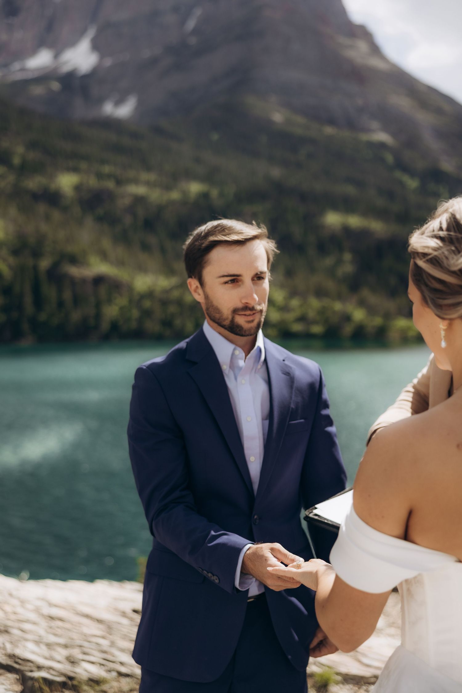 Groom in navy suit holds bride's hands during outdoor wedding ceremony by a turquoise mountain lake.